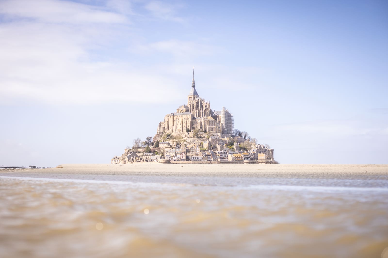Mont Saint-Michel island abbey reflected in shallow tidal waters under a bright blue sky.
