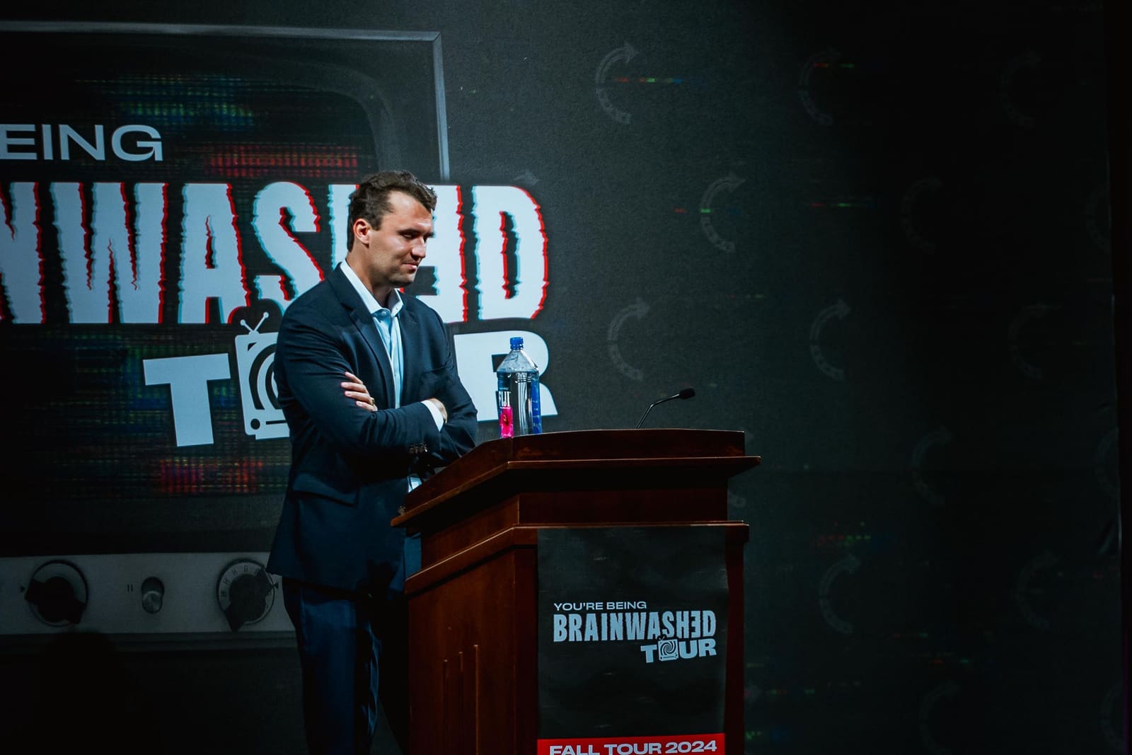 Charlie Kirk at the Brainwashed Tour podium with arms crossed, fall 2024