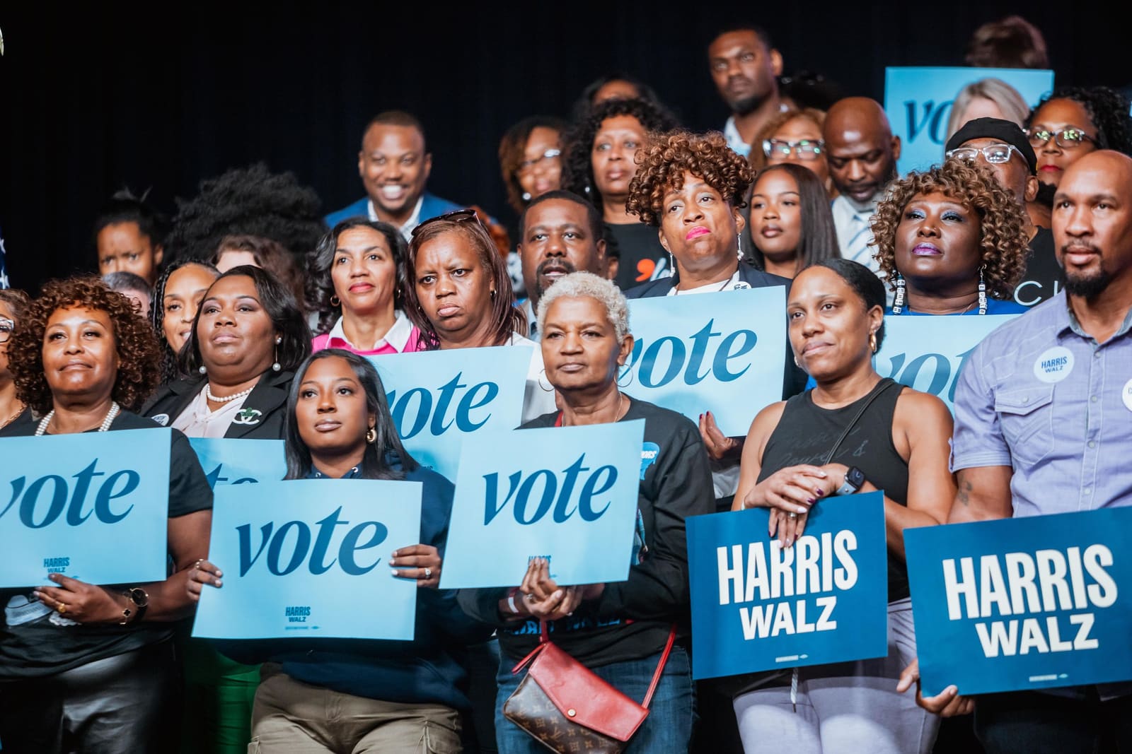 Supporters hold Harris campaign and vote signs at a Kamala Harris campaign rally with a diverse crowd.