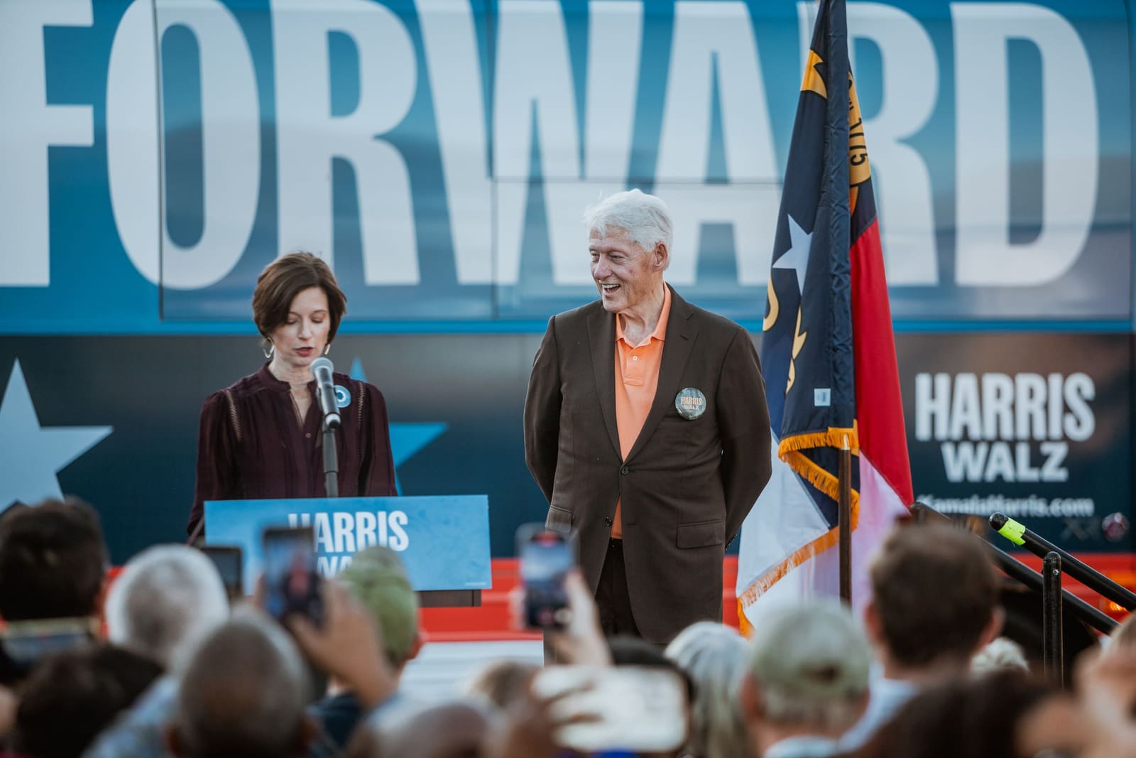 Bill Clinton and another speaker on stage beside the North Carolina flag