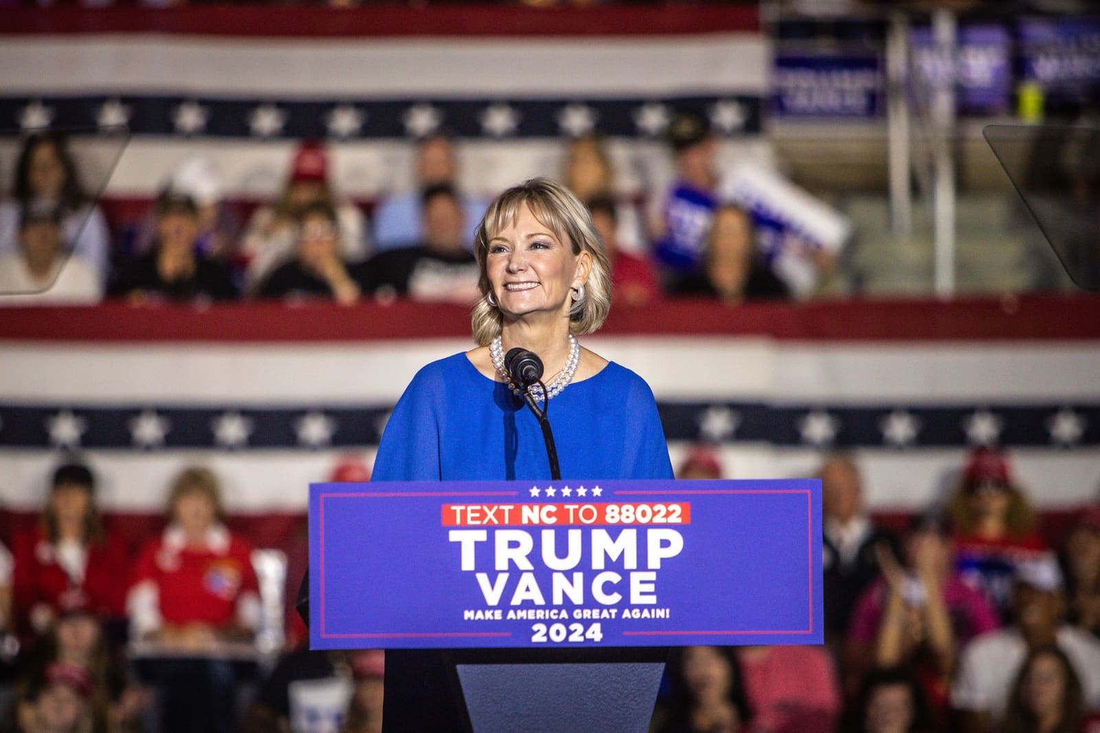 Woman in blue dress speaks at Trump Vance 2024 campaign rally before large crowd.