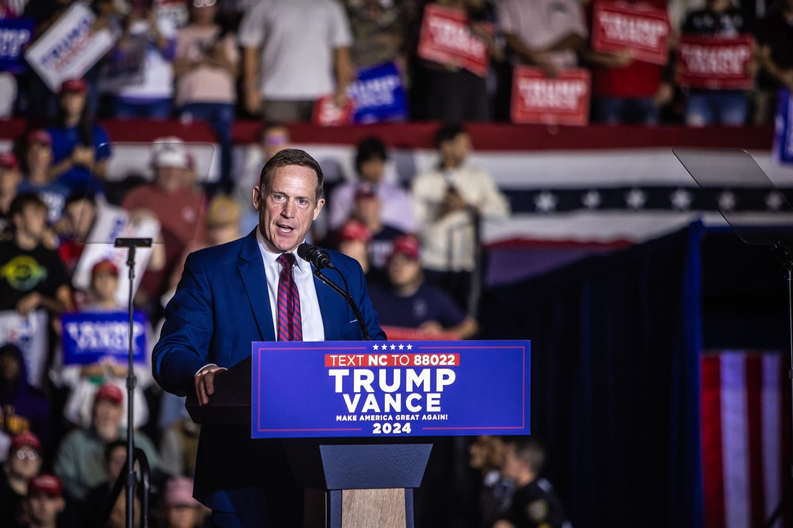 A speaker in a blue suit addresses a large crowd at a Trump Vance 2024 campaign rally in North Carolina.