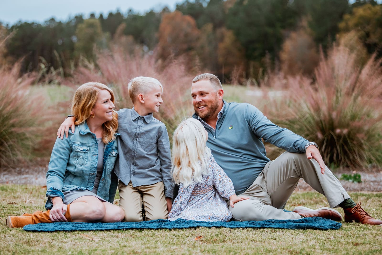 Happy family of four sits on blanket in field, laughing and enjoying outdoor autumn photo session.