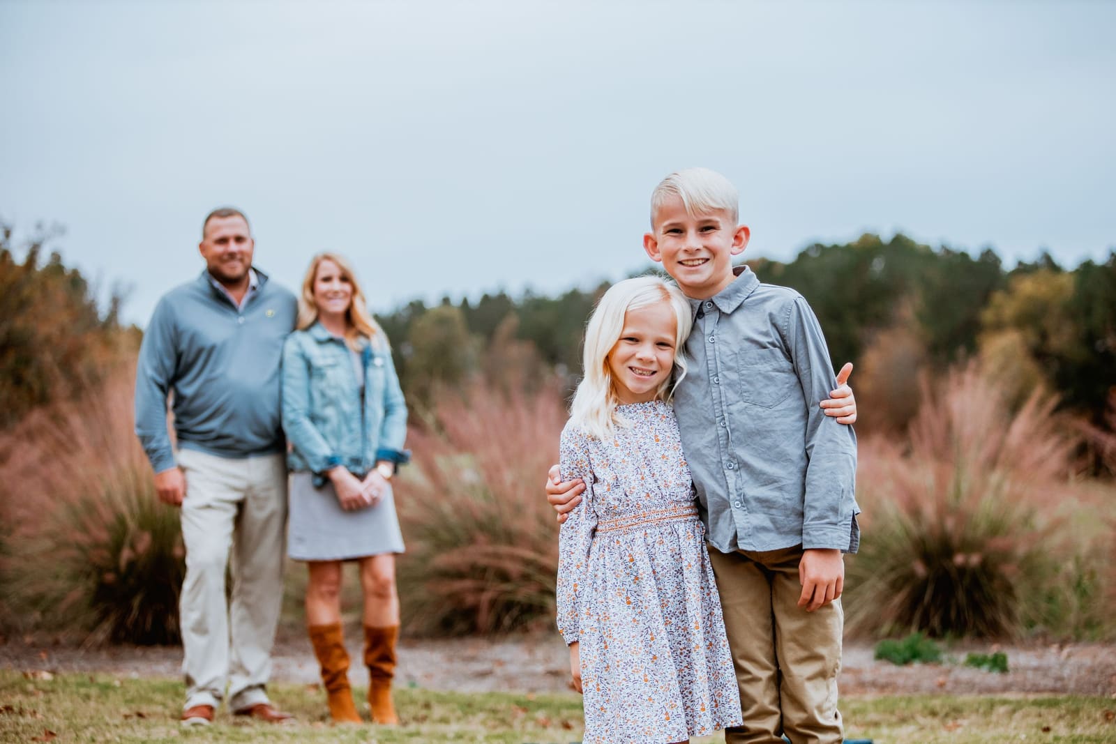 Smiling siblings stand together for family portrait with parents blurred in background amid fall foliage.
