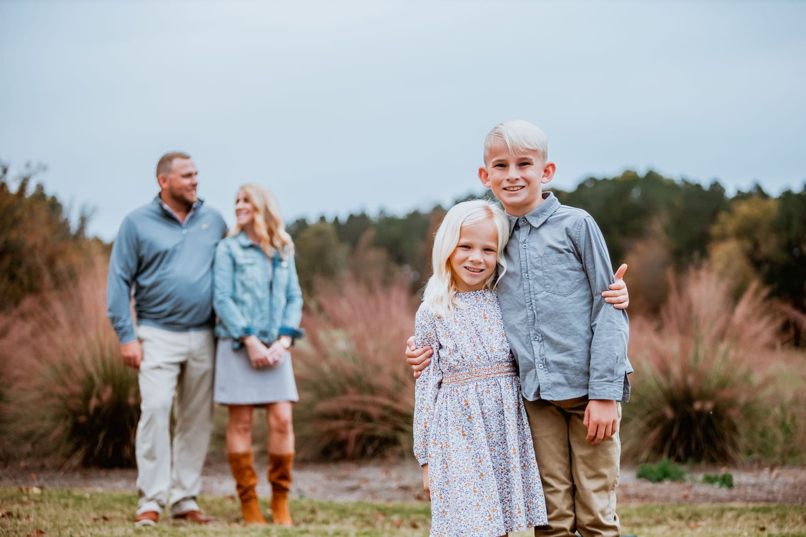 Family of four poses outdoors with children in foreground and parents standing behind among autumn grasses.
