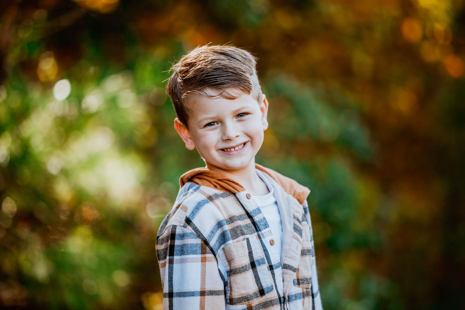Cheerful young boy in a plaid shirt smiling warmly against a vibrant autumn bokeh background.