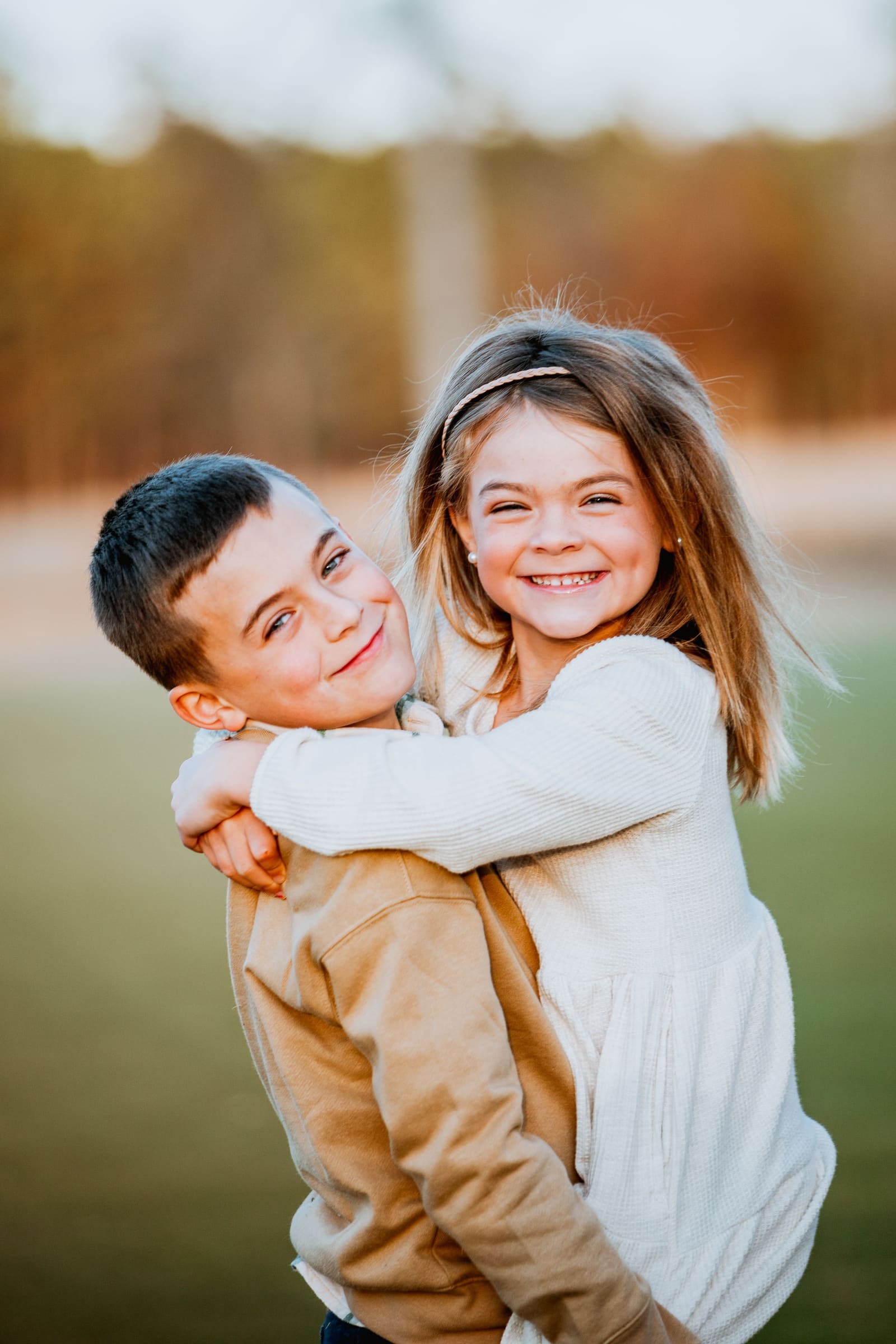 Two happy children hugging outdoors, girl smiling brightly while boy gives her a piggyback in golden afternoon light.