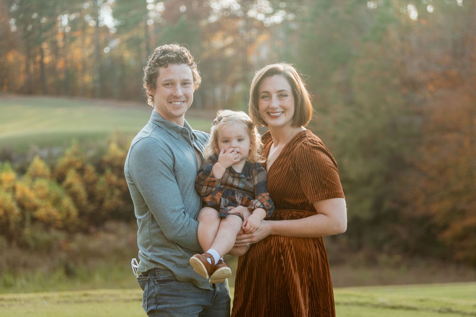 Smiling family of three with dad holding toddler daughter stands in golden autumn field at sunset.