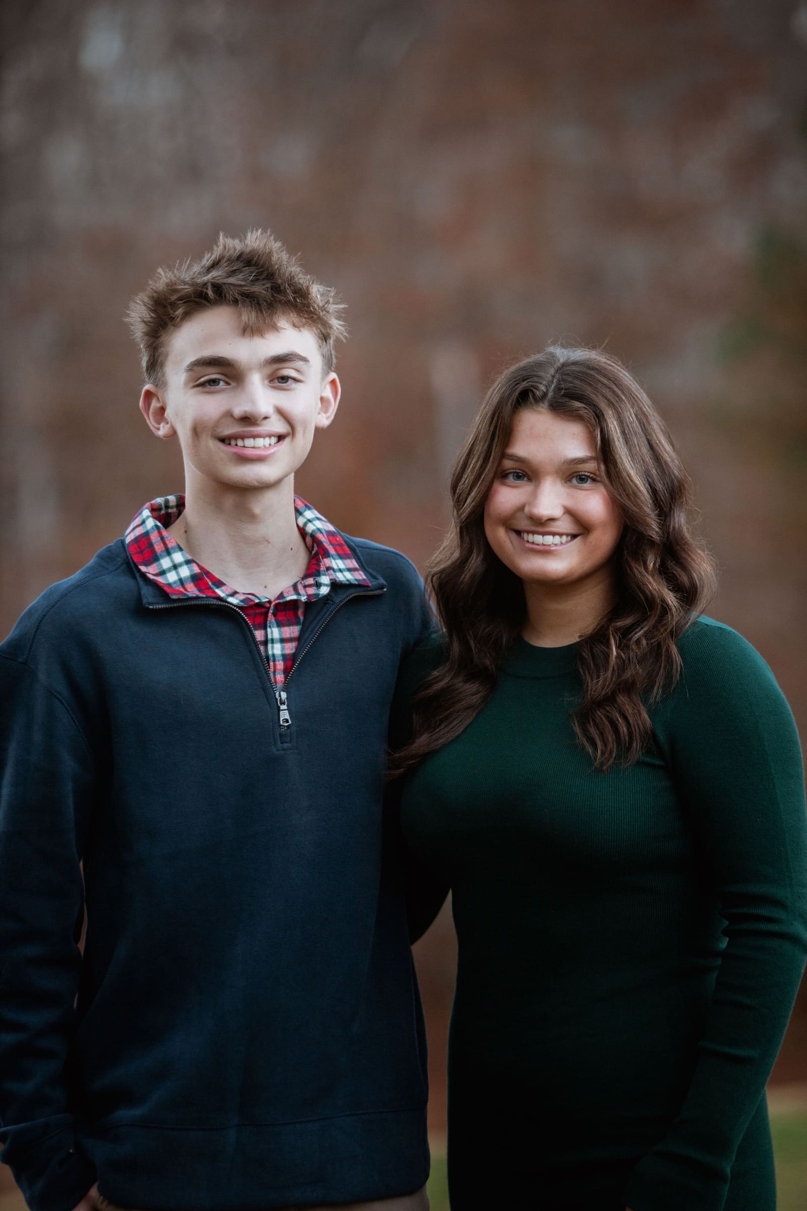 Teen boy in navy pullover and girl in green dress smile together in front of a brick wall.