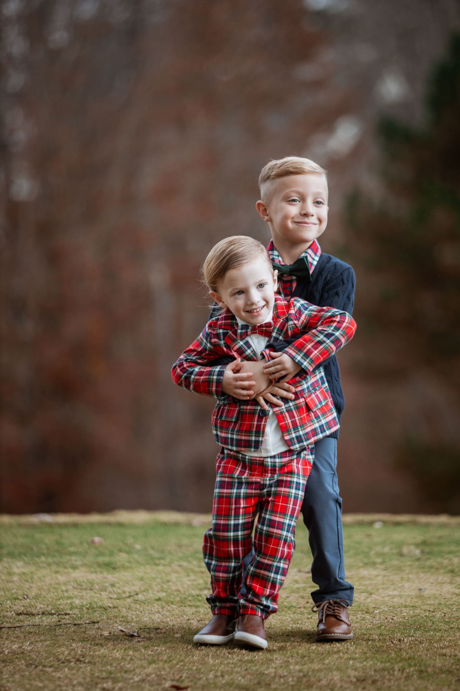 Older boy in navy hugs younger toddler in red plaid outfit on a grassy outdoor setting.
