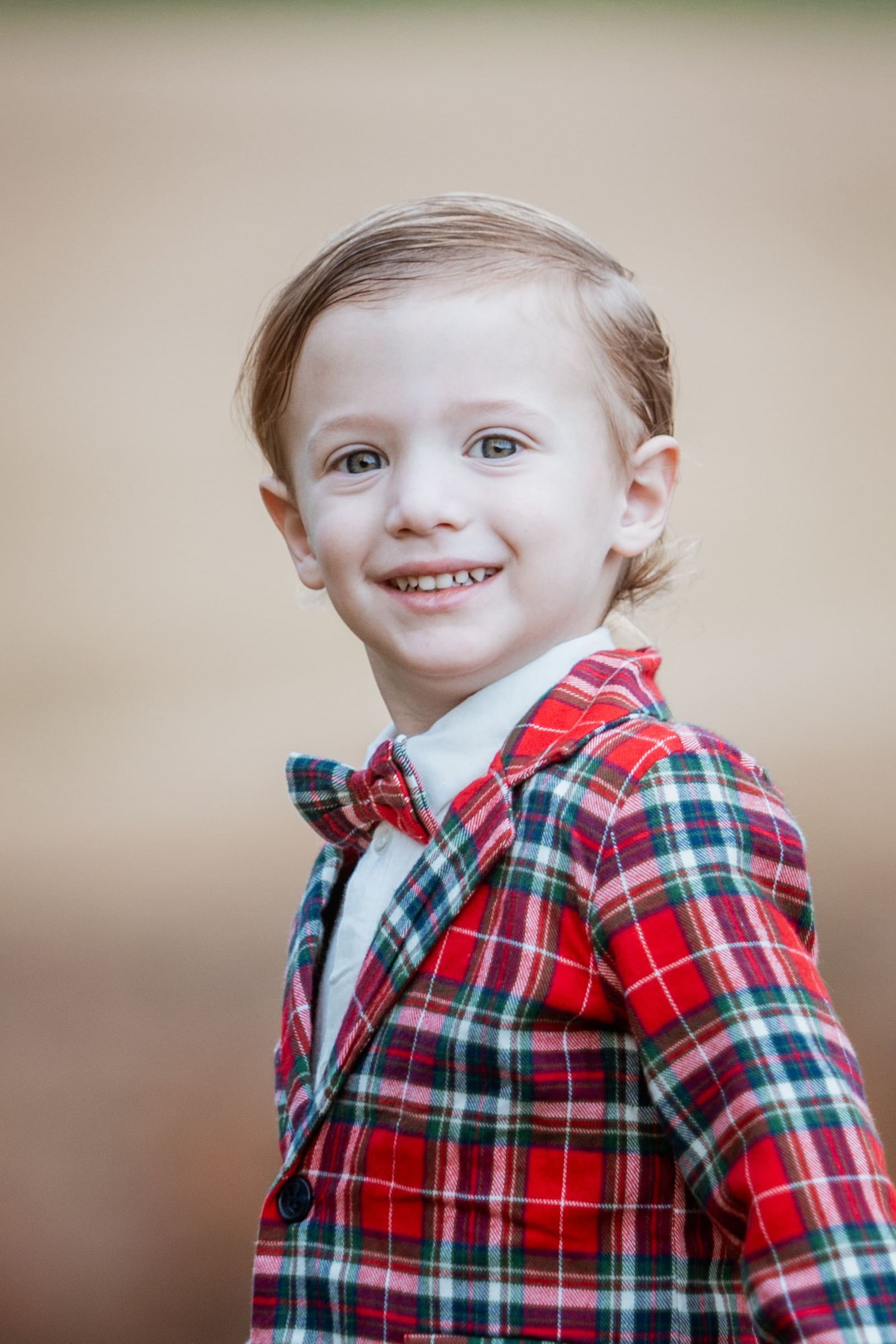 Close-up portrait of smiling young boy in red plaid jacket and bow tie outdoors.