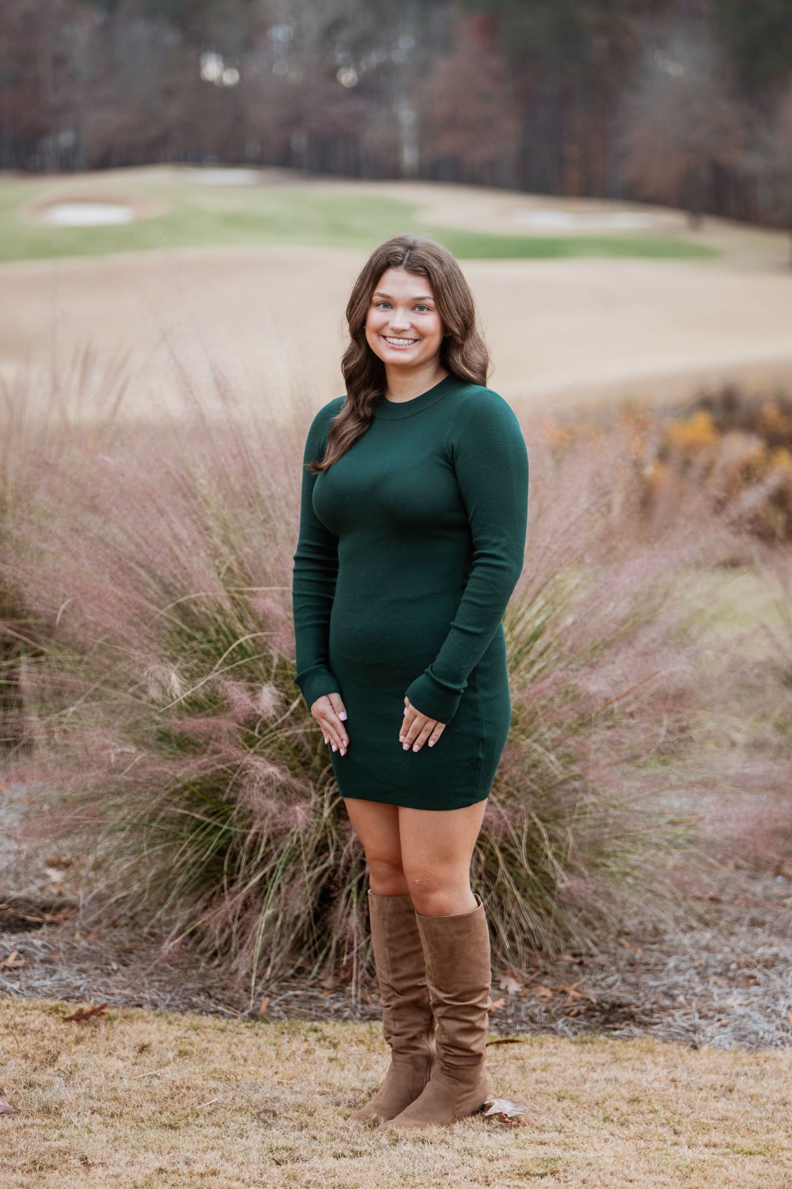 Young woman in green long-sleeve mini dress and brown boots on golf course.