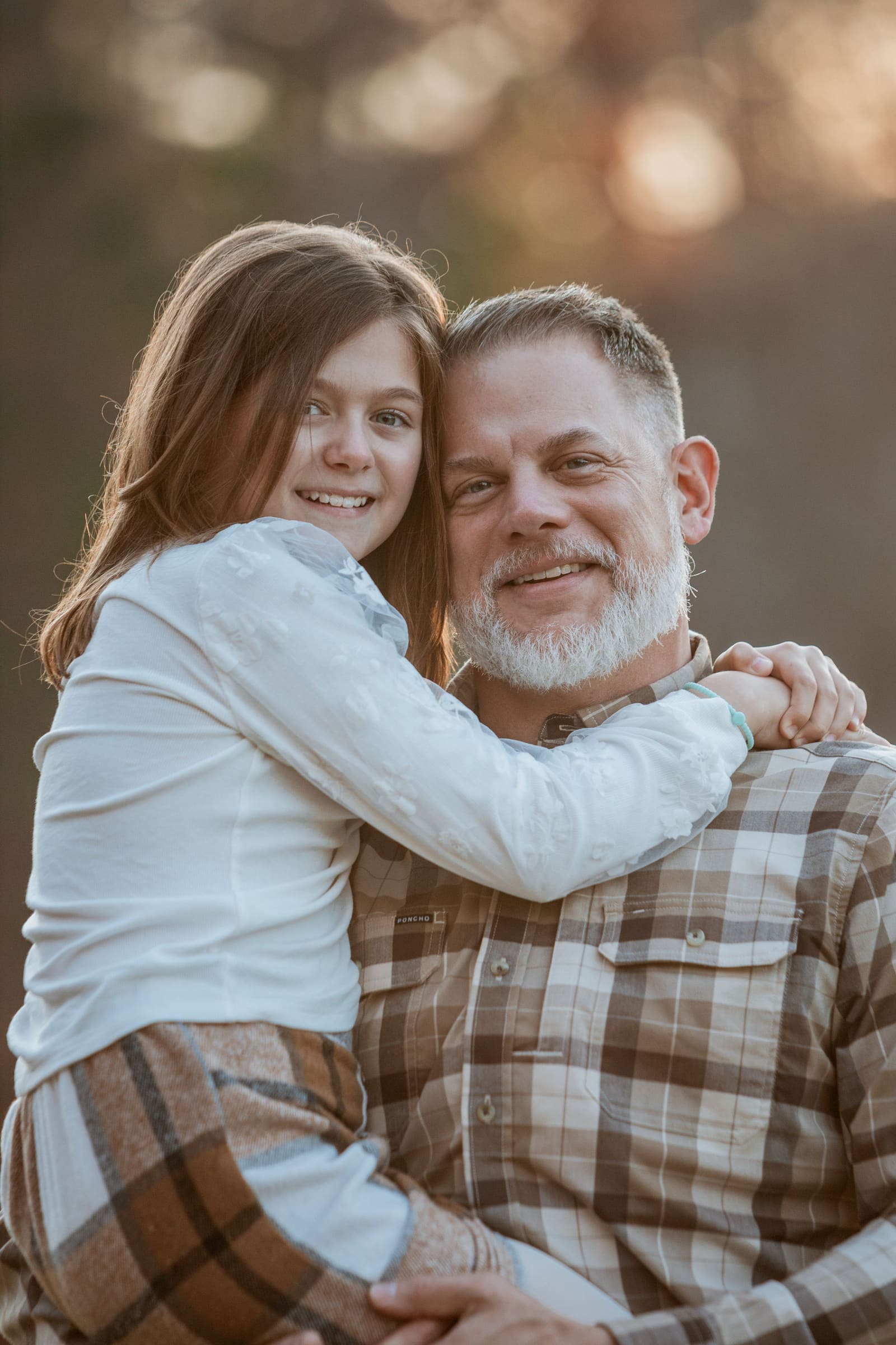 Smiling girl hugging bearded father from behind in warm outdoor autumn light.