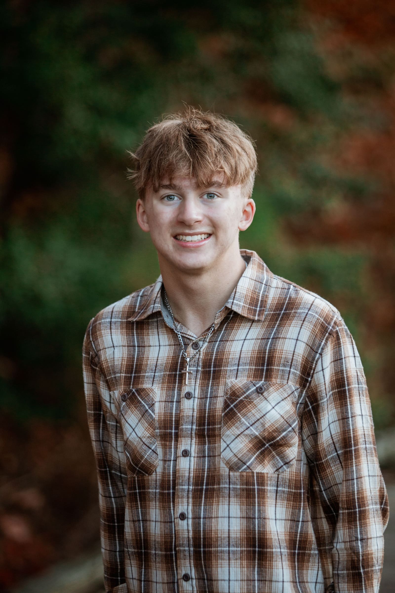 Smiling teenage boy in brown plaid shirt posing outdoors with colorful autumn foliage behind him.