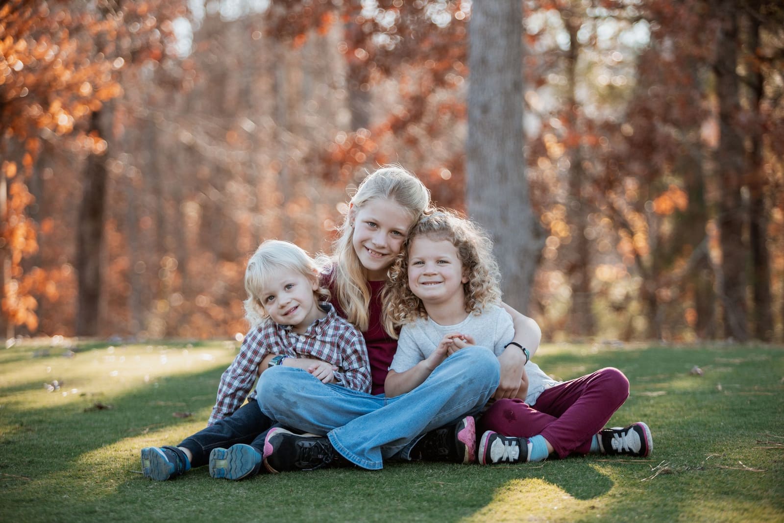 A smiling blonde mother sits on grass hugging her two young children with fall foliage backdrop.