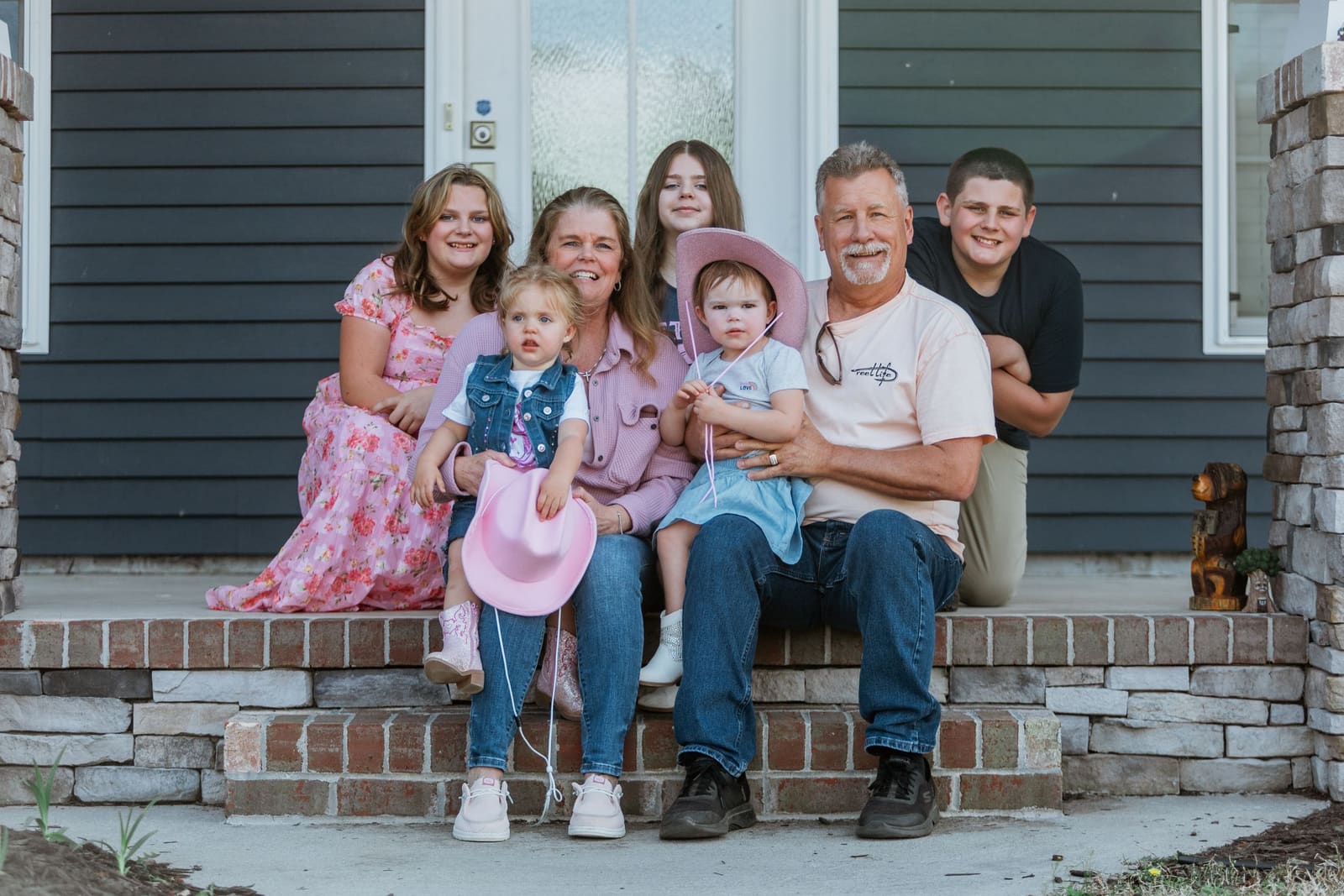 A smiling family of six poses together on brick front porch steps of a gray house.