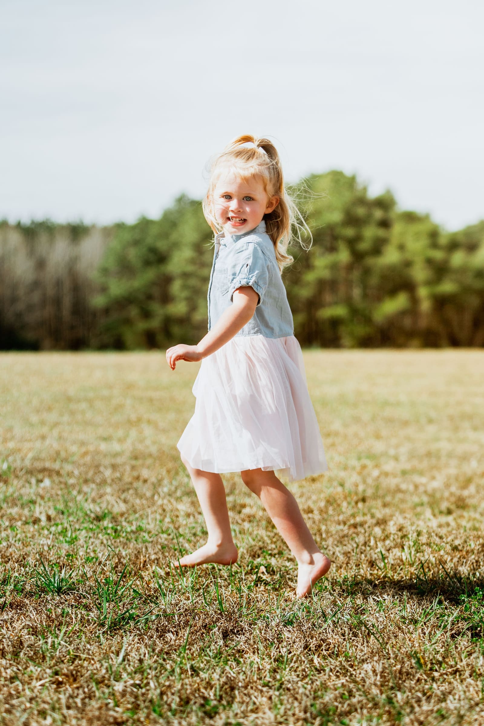 A happy blonde girl in a blue top and white tulle skirt runs barefoot across a sunny green field.