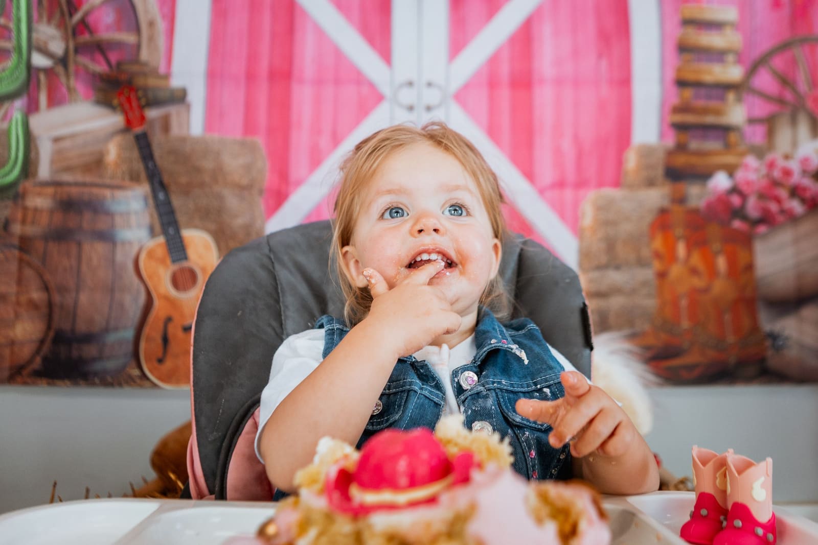 Toddler girl in denim vest eating cake at a pink barn-themed birthday party.