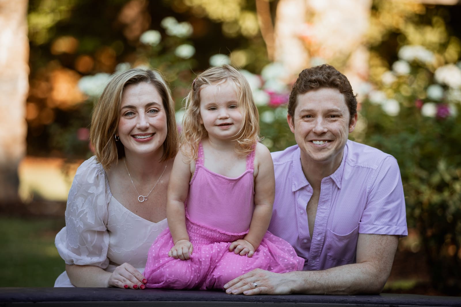 Smiling family of three posing outdoors with bokeh floral background, parents flanking happy toddler.