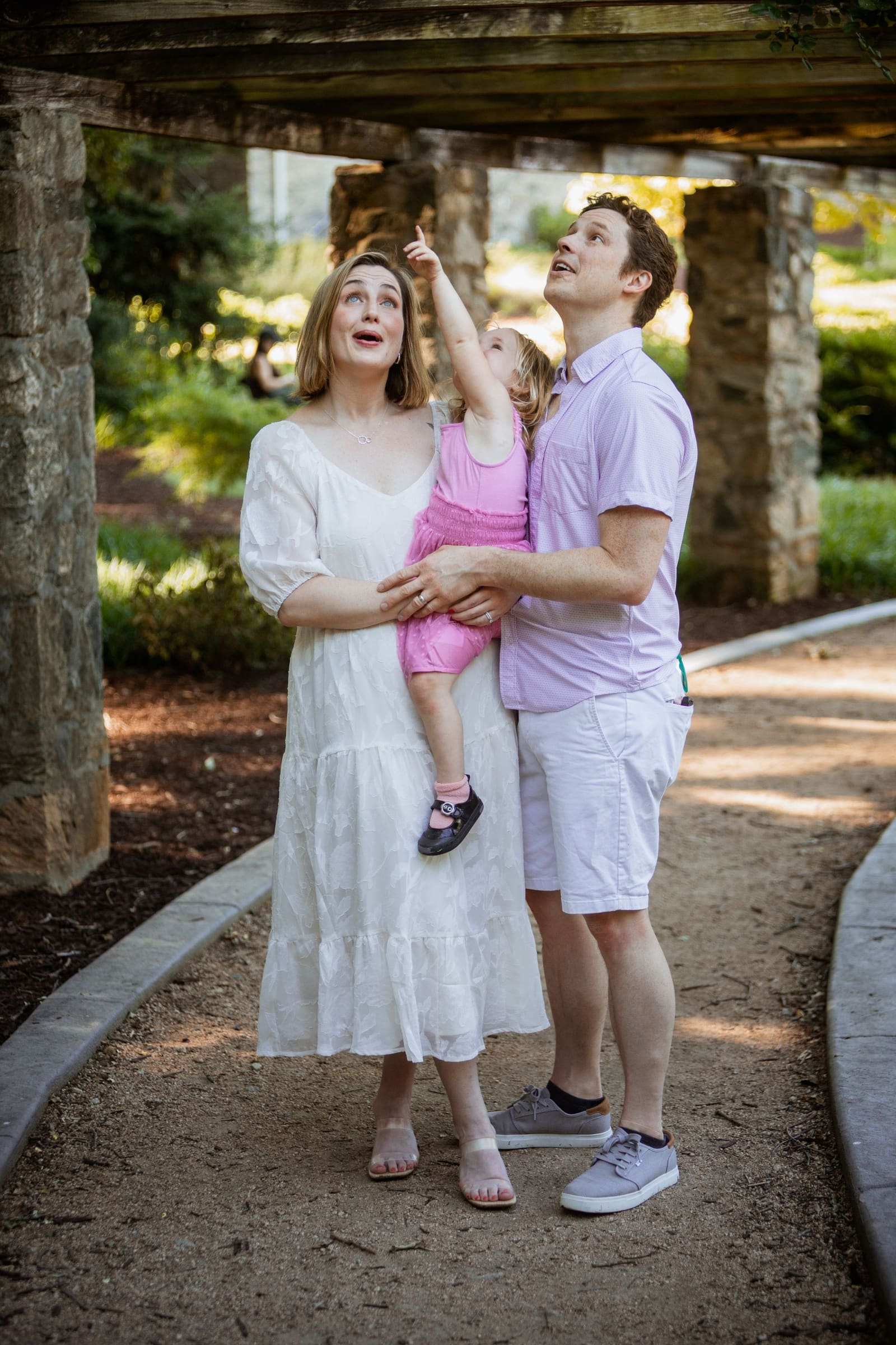 Family of three looking up with curiosity as toddler points skyward under a rustic stone pergola.