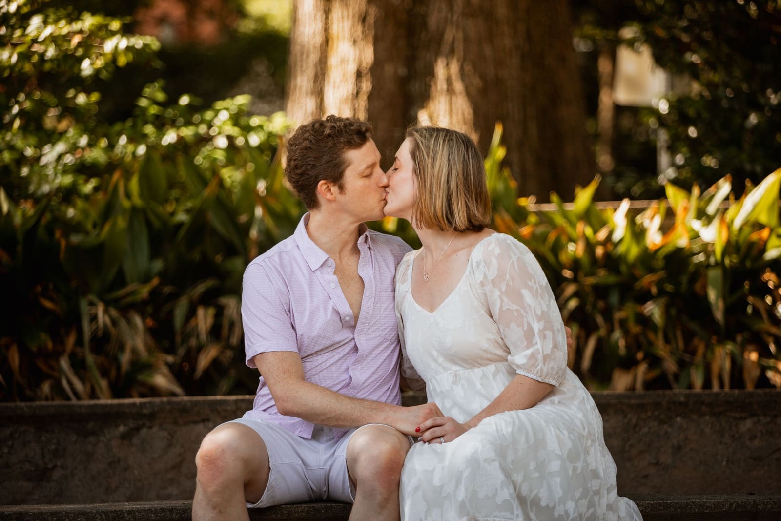 Couple sharing a romantic kiss while seated on stone steps surrounded by lush greenery.