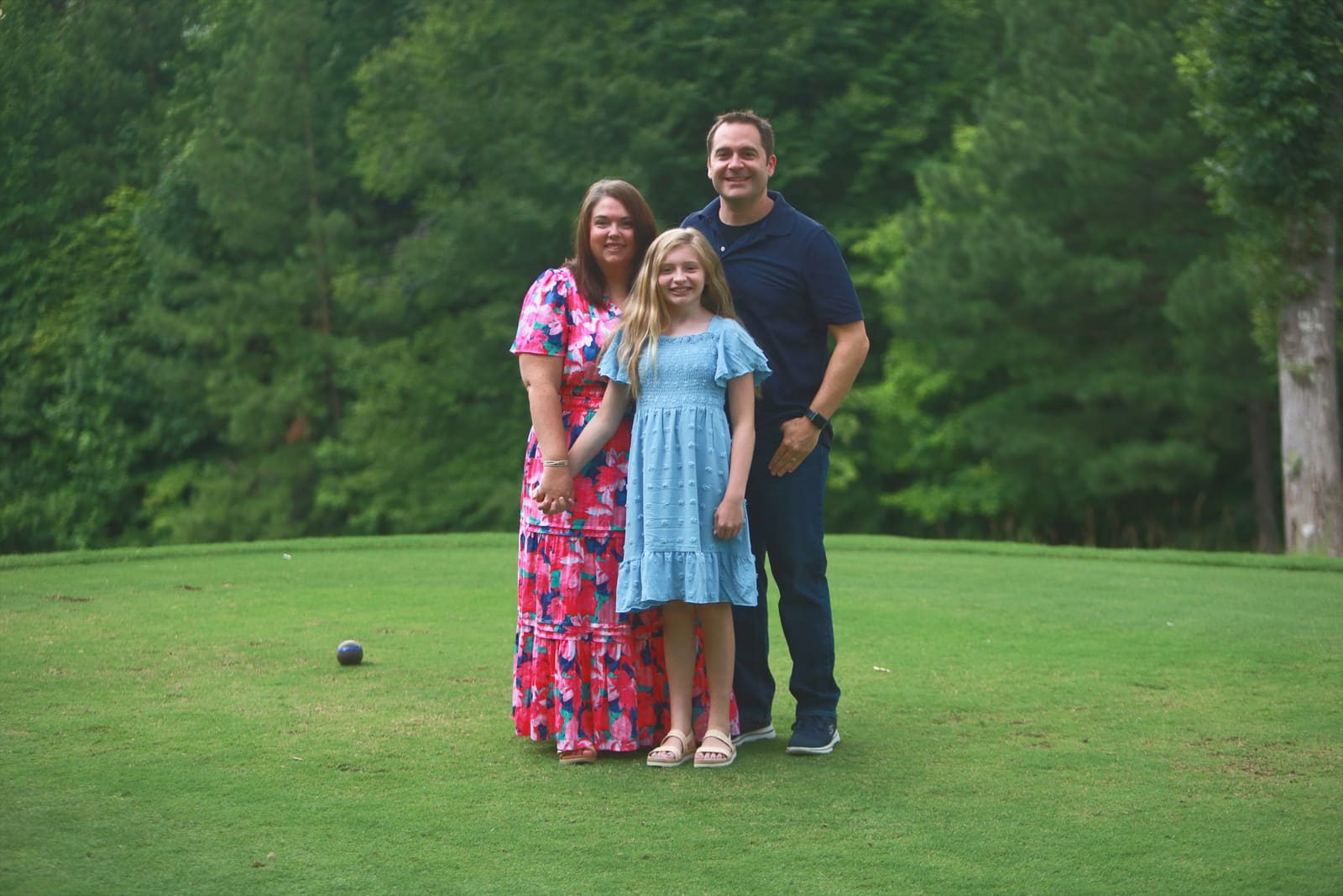 Parents and daughter standing together on a green golf course lawn with tall pines behind them