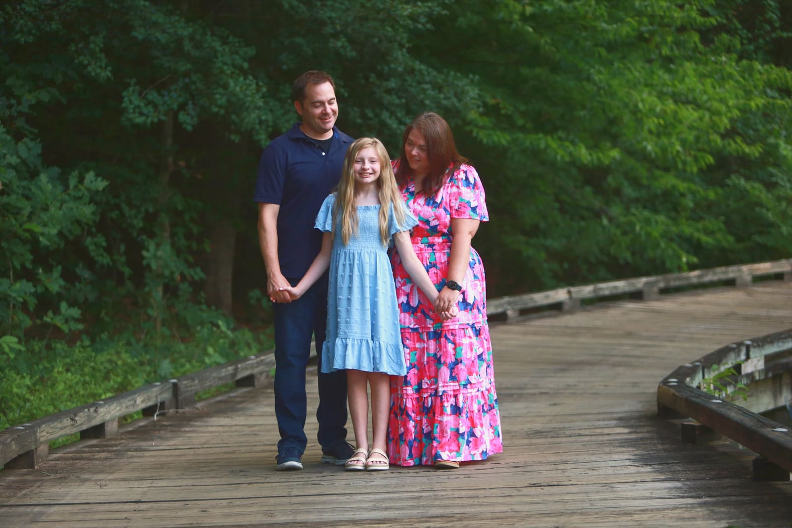 Mother, father and daughter holding hands on a wooden boardwalk in a green wooded park