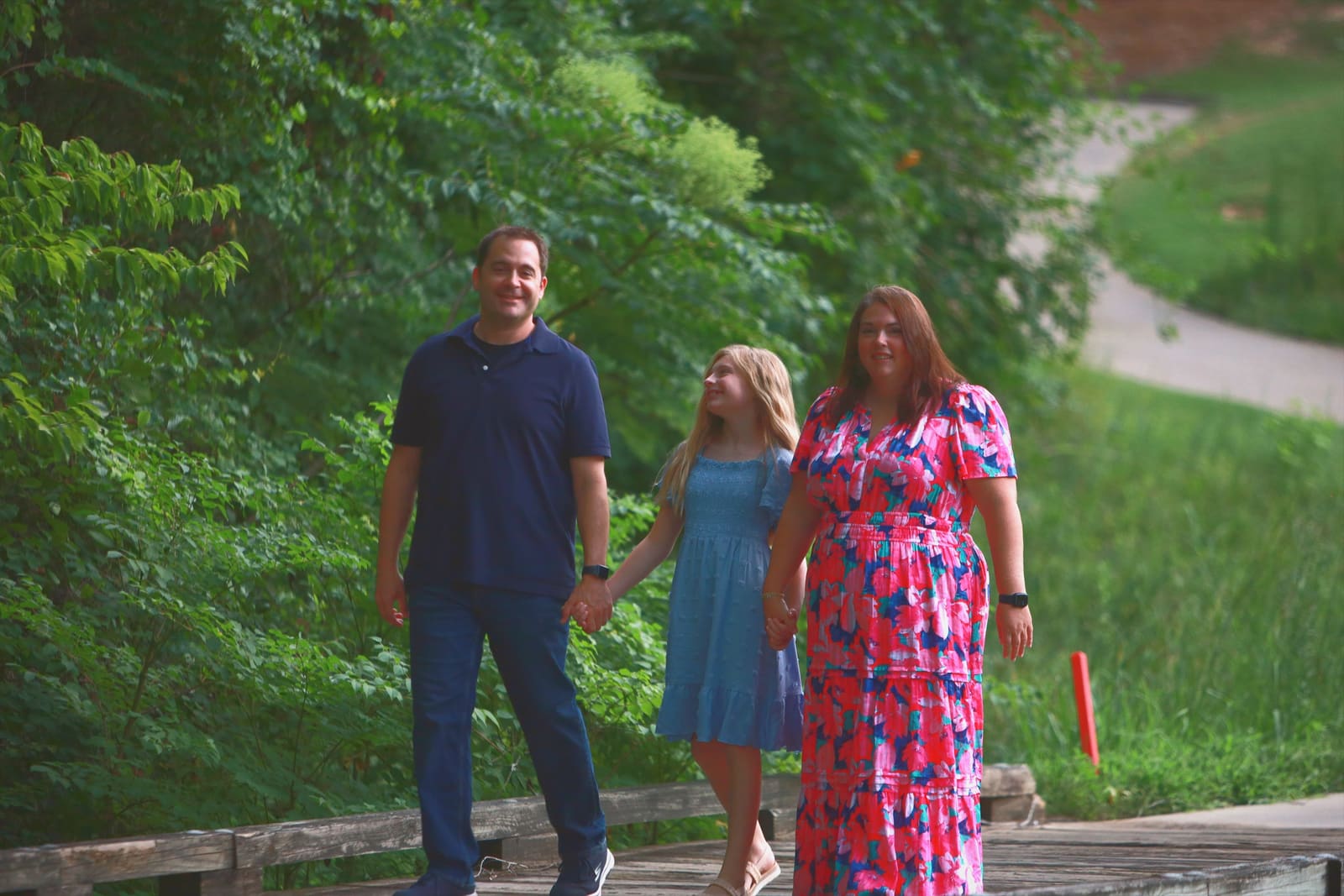 A family walking hand in hand along a park boardwalk surrounded by summer greenery