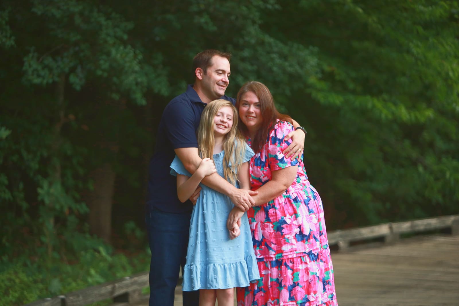 Parents and daughter in a close family hug on a boardwalk with deep green trees behind