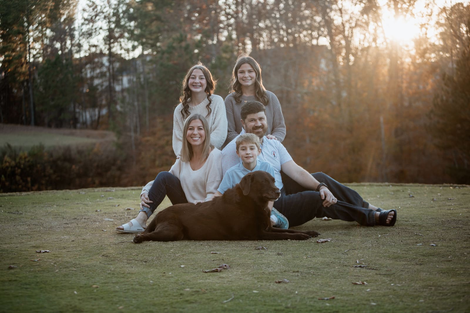 A family seated together on the grass with their dog as the sun sets behind autumn trees