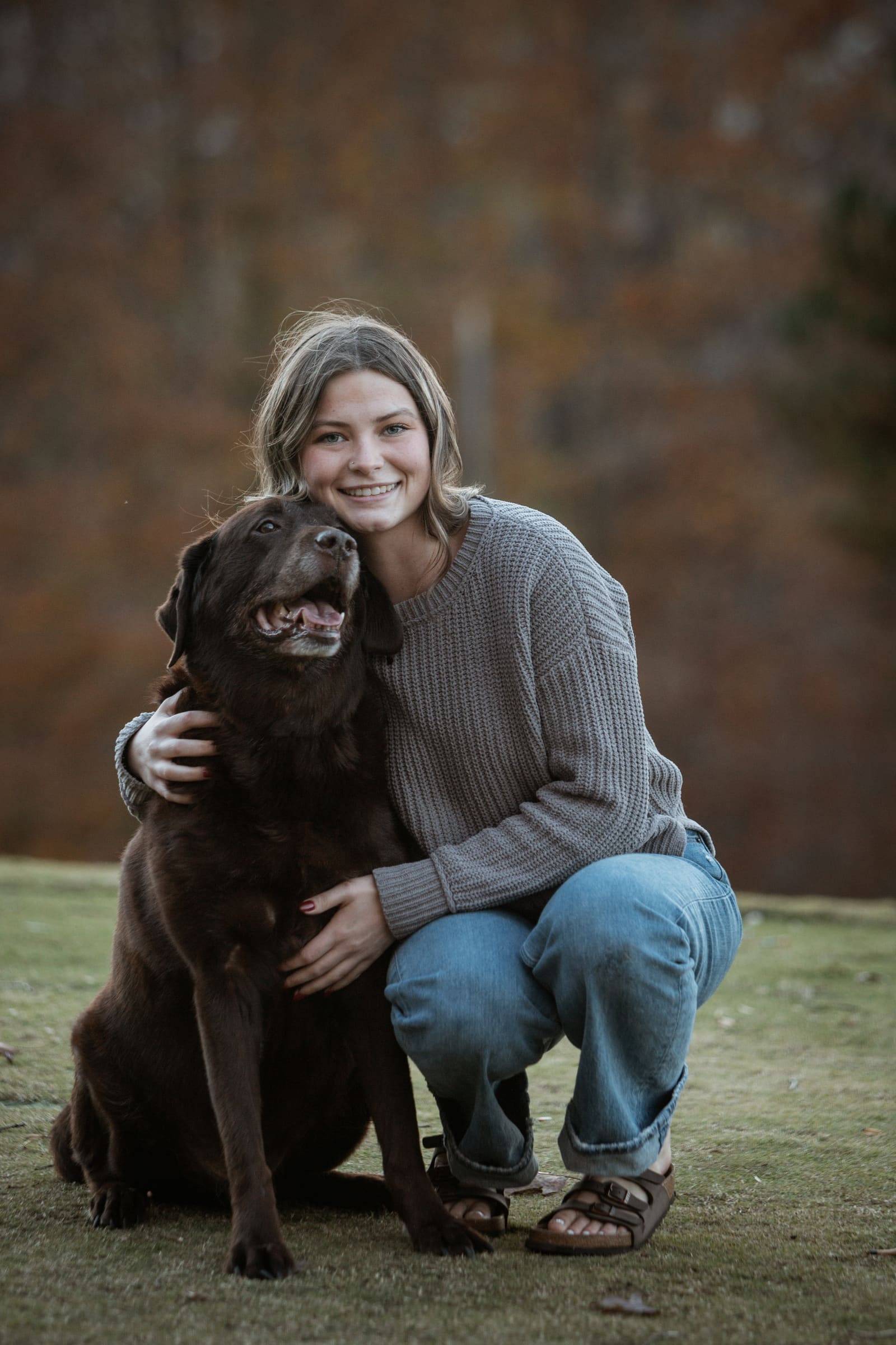 A teenage girl crouching to hug her chocolate Labrador in warm autumn light
