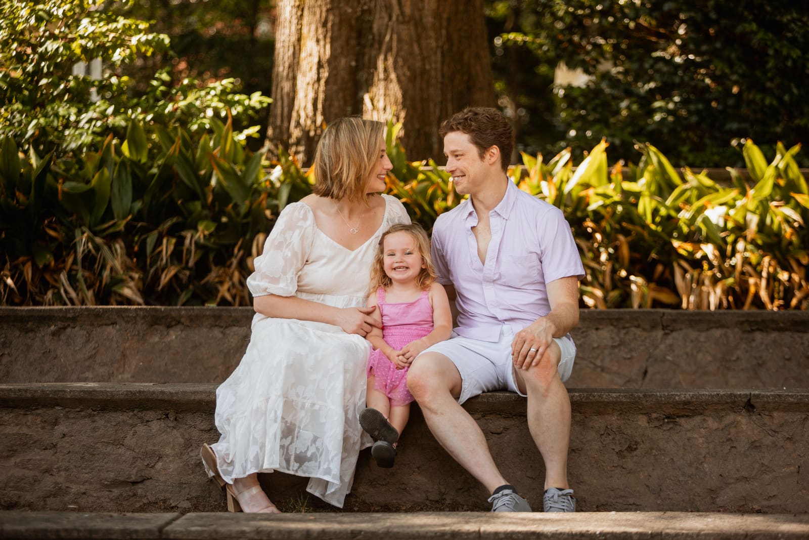 A family of three sitting on a stone ledge in a leafy garden