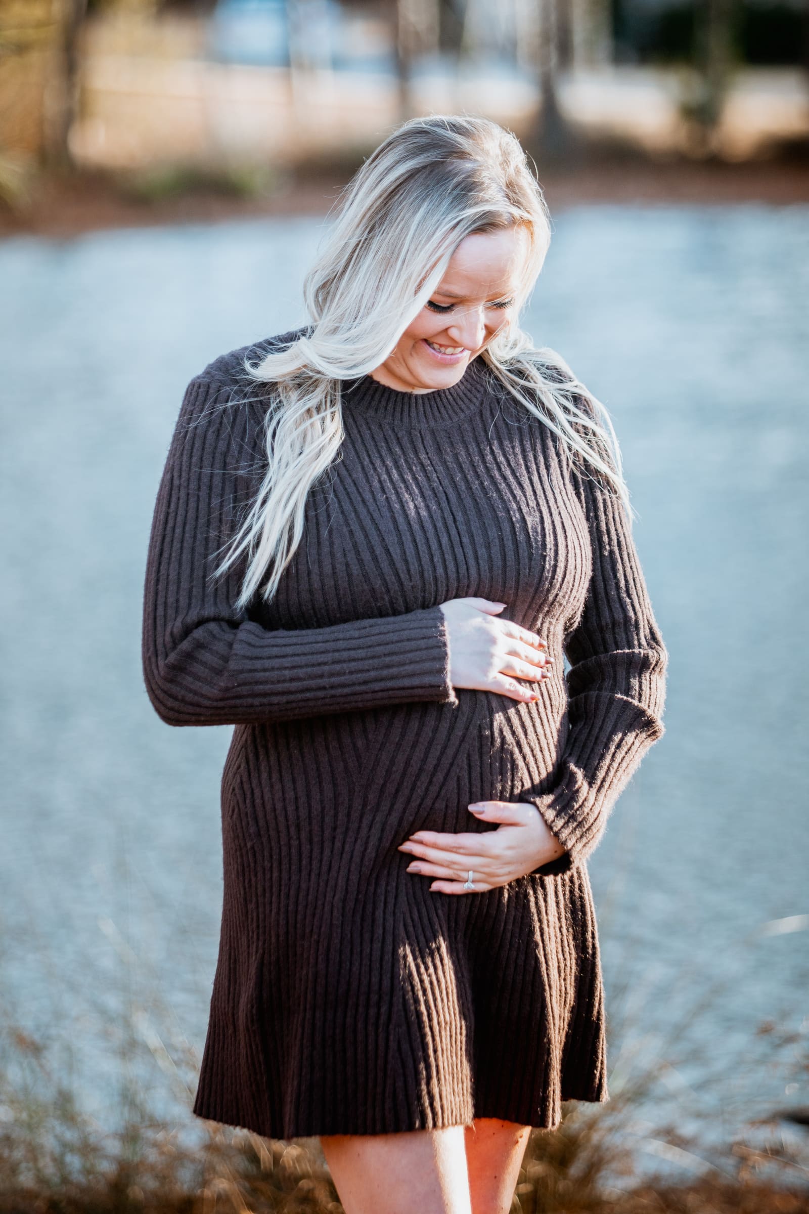 An expecting mother looking down at her bump by a lake with a soft blurred background