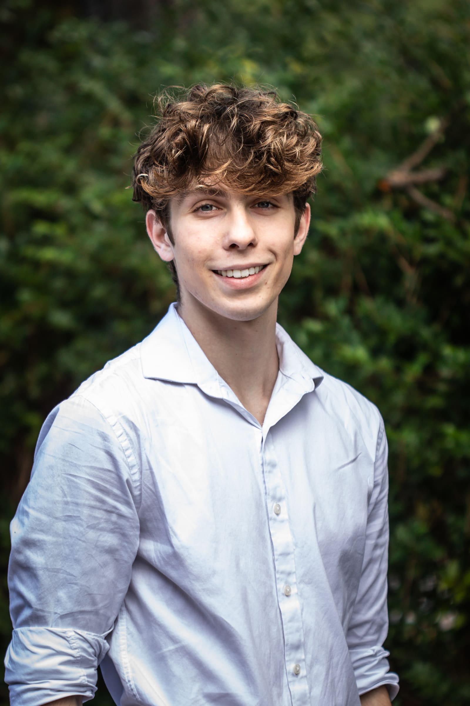 Gabe Key smiling in a white shirt, green leaves behind him