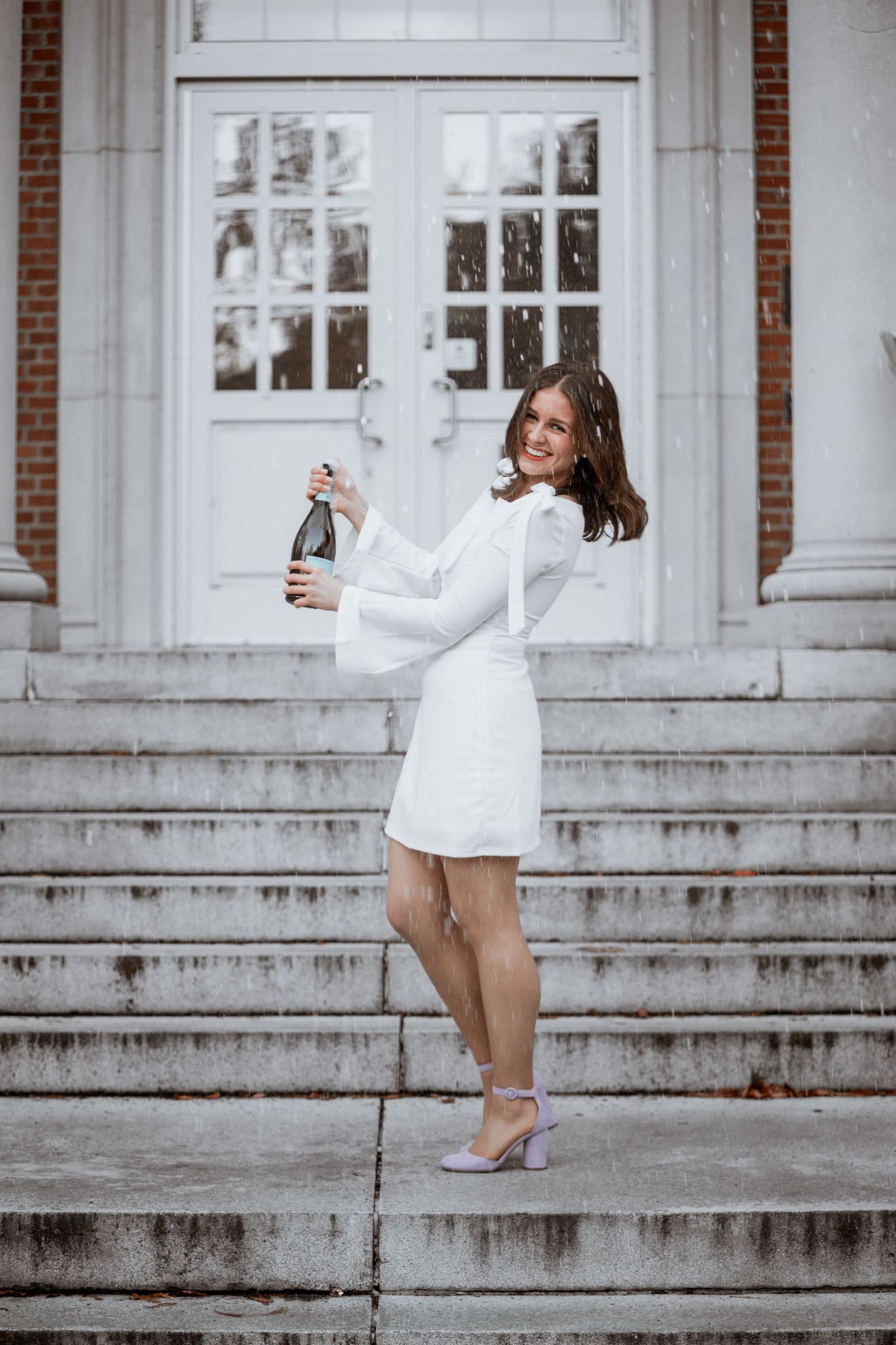 Woman in white mini dress popping champagne bottle on stone steps outside elegant building.