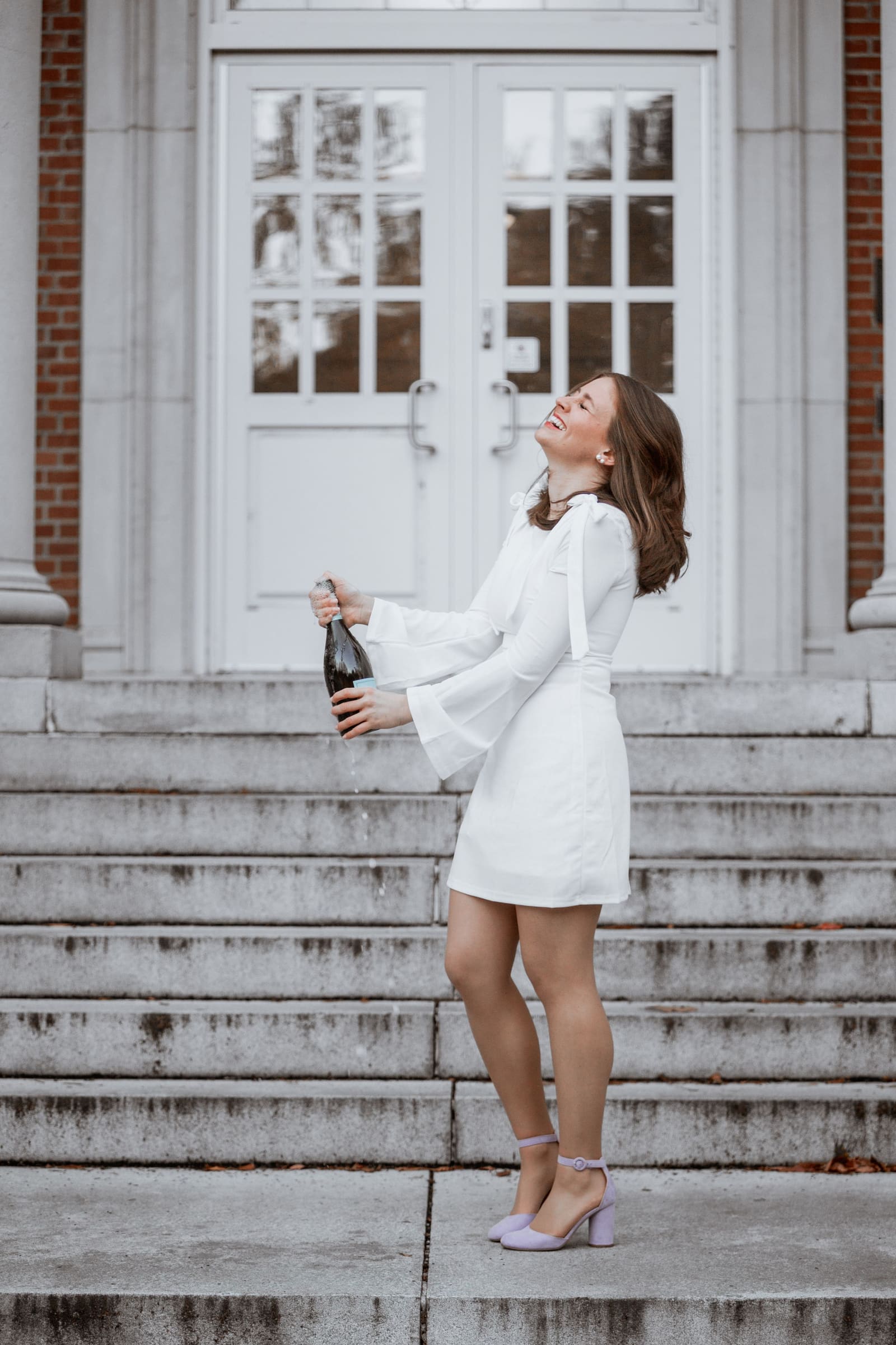 Young woman in white dress celebrates graduation by popping champagne bottle on brick building steps.