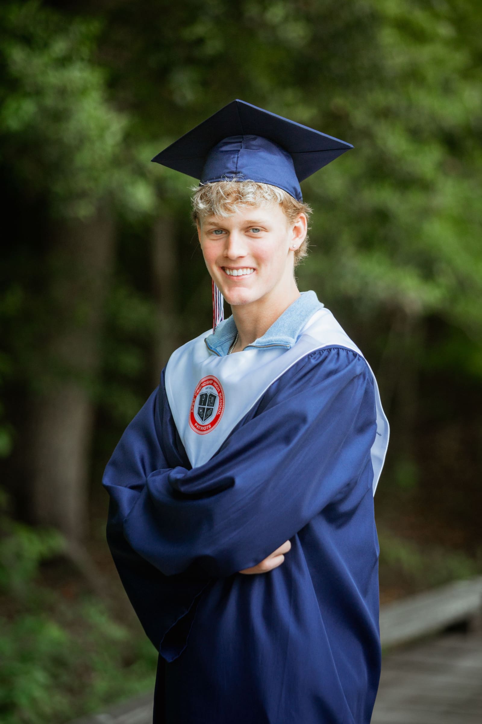 Smiling graduate poses in blue cap and gown with arms crossed on outdoor wooden boardwalk path.