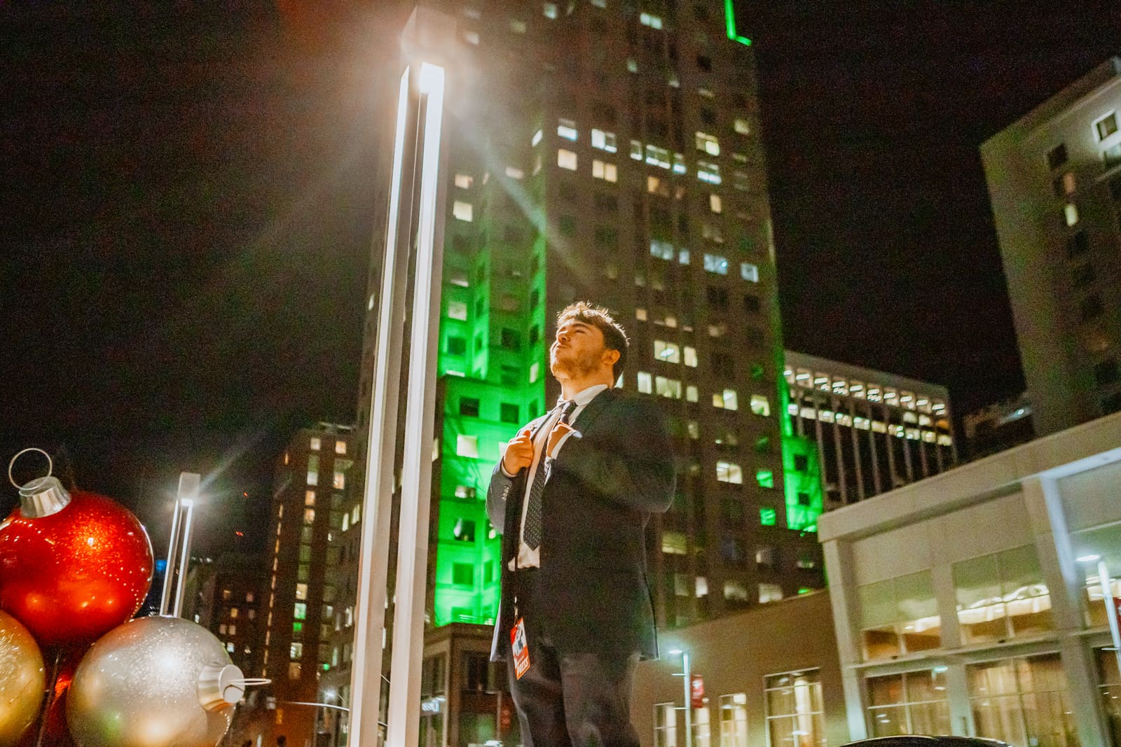 A man looking up at a lit green skyscraper at night beside large holiday ornaments