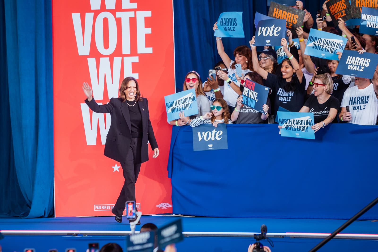Kamala Harris walking on stage in front of a red We Vote We Win banner