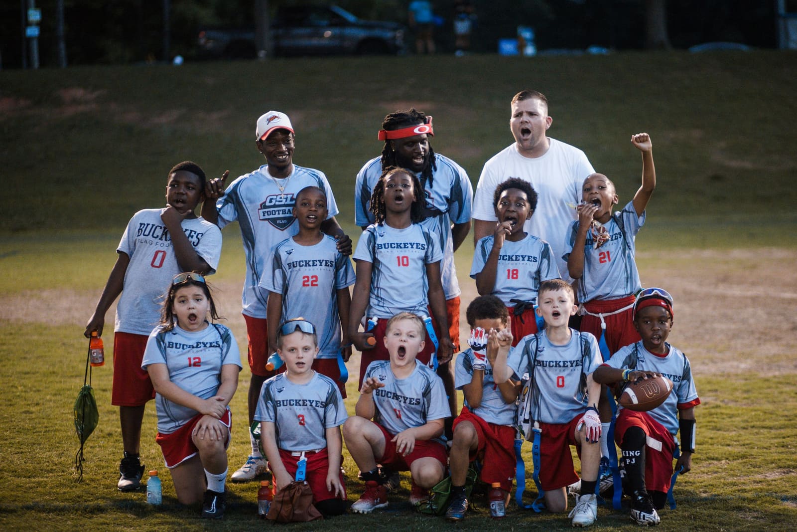 Youth flag football team in blue uniforms celebrating with coaches on a grass field at night.