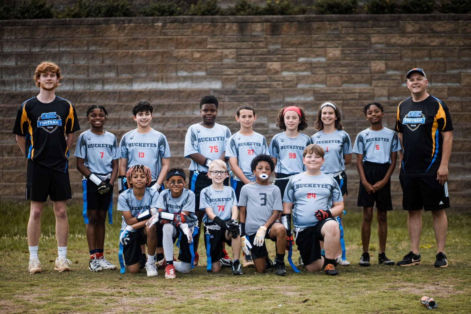 Youth flag football team in gray uniforms posing with two coaches against a brick wall.