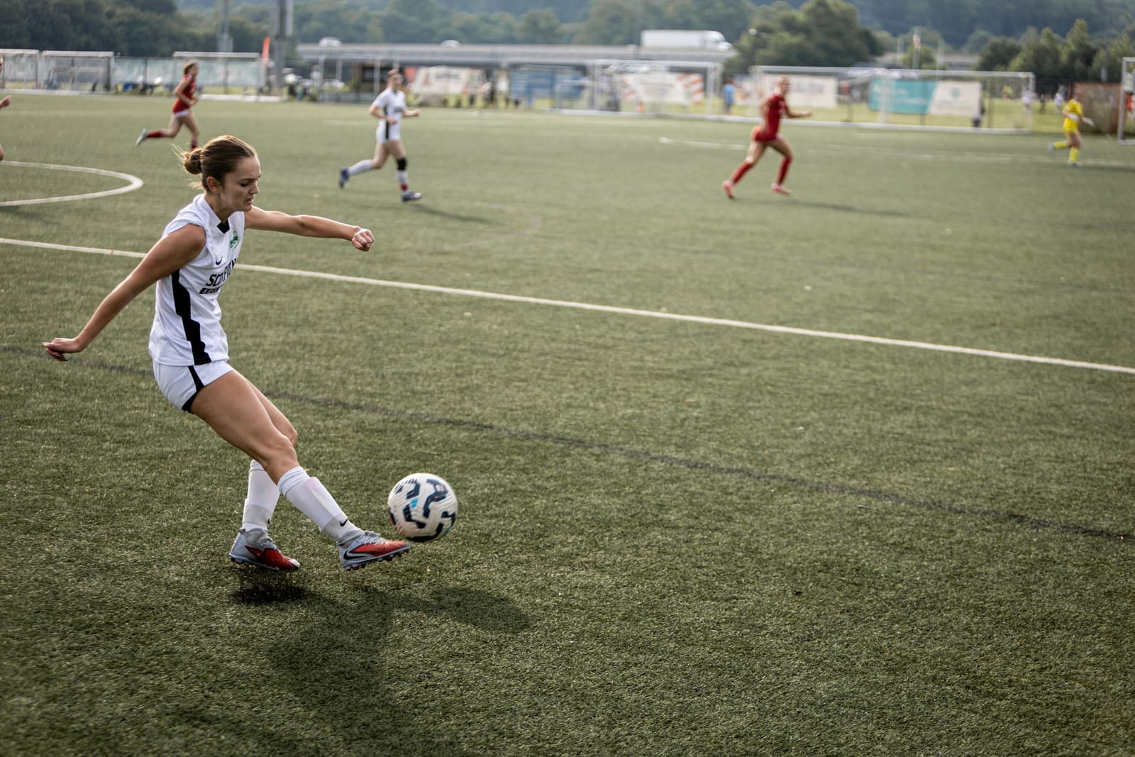 A soccer player in white striking the ball with a full follow-through on a green pitch